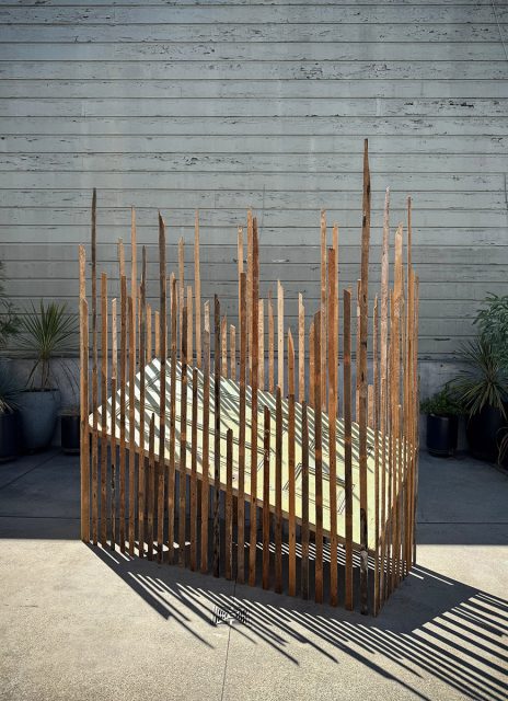 A geometric wooden sculpture with uneven vertical slats stands on a concrete patio against a weathered wall, with potted plants in the background.