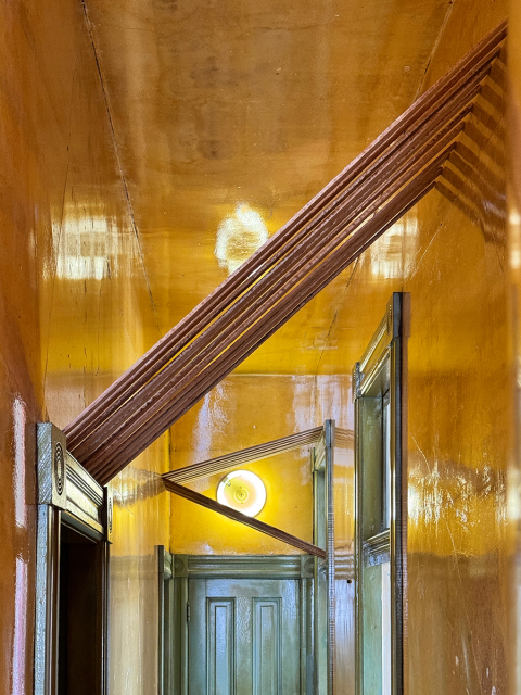 Narrow hallway with glossy yellow walls, angled brown beams crossing overhead, and a round light fixture near green doors at the end.