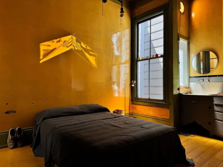 A small room with yellow walls, a bed covered in black bedding, a window with daylight, a mirror, a sink, and a pair of shoes on the floor.