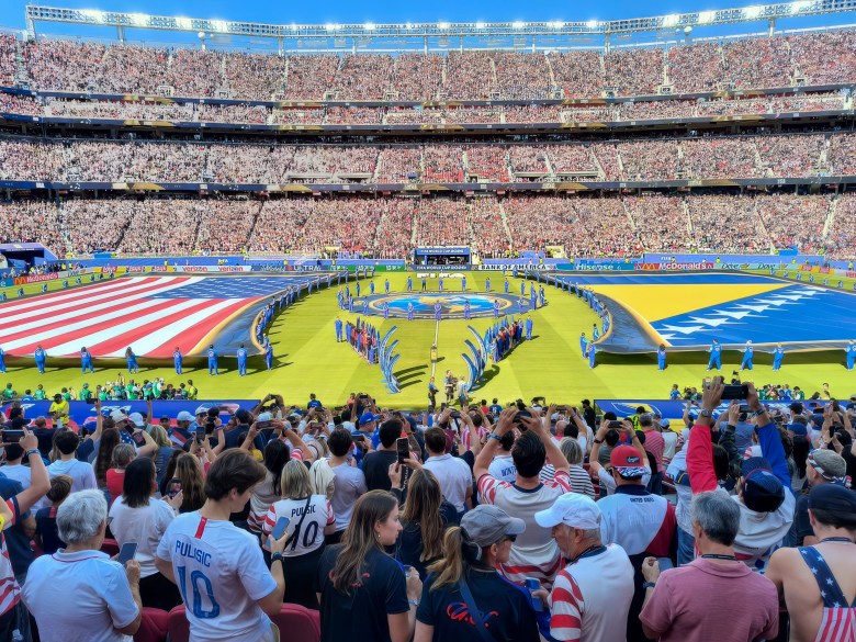 A large stadium filled with spectators watches a ceremonial display featuring giant flags of the United States and Bosnia and Herzegovina on the field.