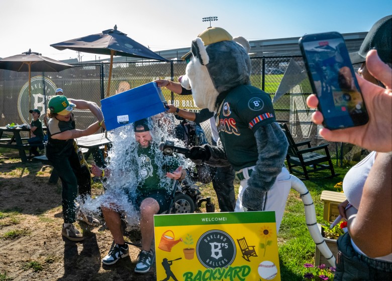 A person in a green shirt is drenched as a mascot pours water from a blue container during an outdoor event.