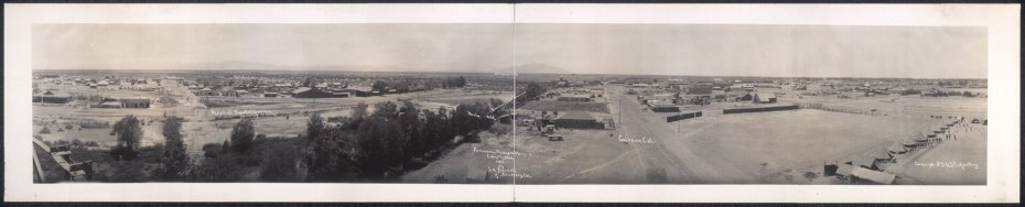 Wide panoramic black-and-white view of a military camp with tents, buildings, open fields, and distant mountains under a clear sky. Handwritten labels mark some locations.