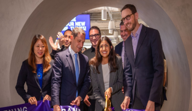 A group of seven well-dressed people smile as a woman cuts a purple ribbon at a grand opening event inside a modern, curved tunnel entrance.
