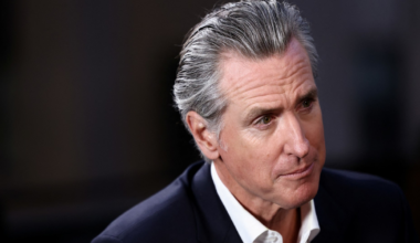 A middle-aged man with slicked-back gray hair wears a dark suit jacket and a white shirt, looking thoughtfully to his right.