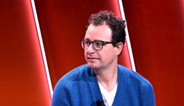 A man with curly dark hair and glasses wears a blue cardigan over a white shirt, sitting against a red background with vertical light strips.