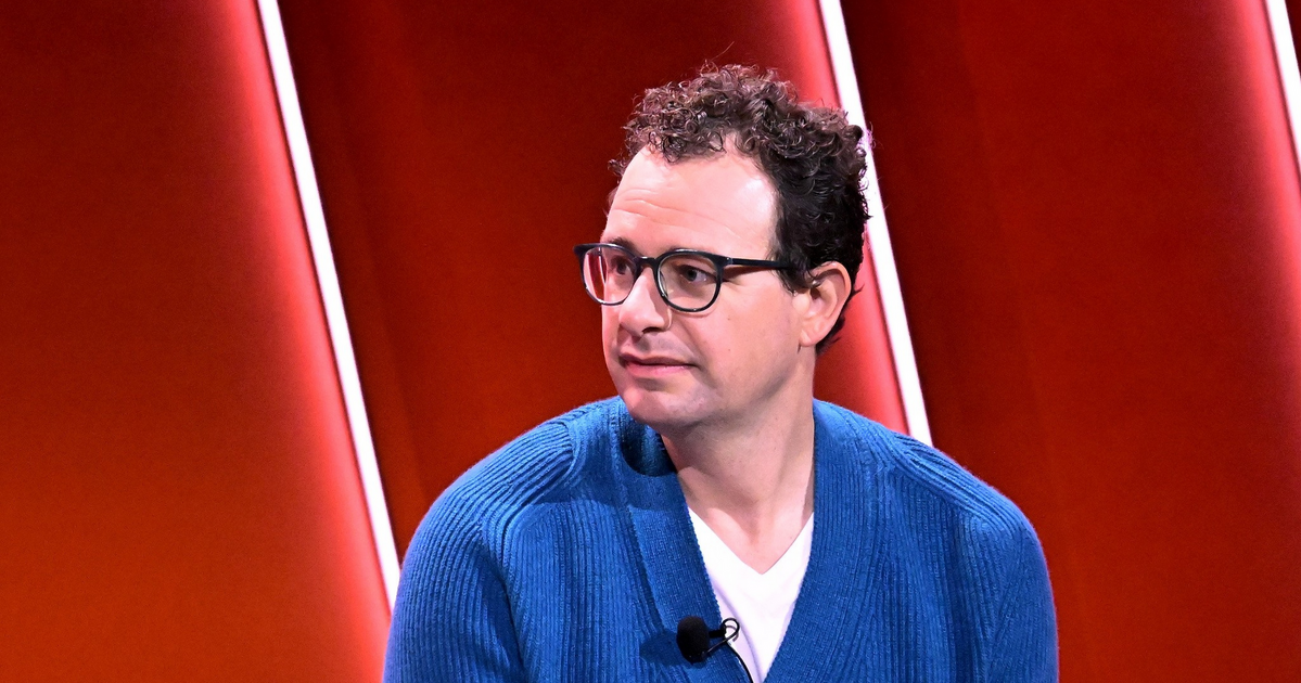 A man with curly dark hair and glasses wears a blue cardigan over a white shirt, sitting against a red background with vertical light strips.