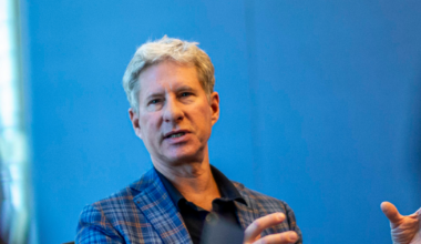 A man with gray hair wearing a blue plaid jacket gestures with his hands while speaking against a blue background.