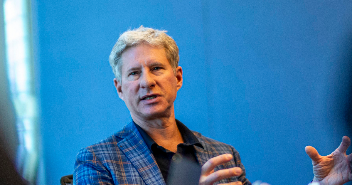 A man with gray hair wearing a blue plaid jacket gestures with his hands while speaking against a blue background.