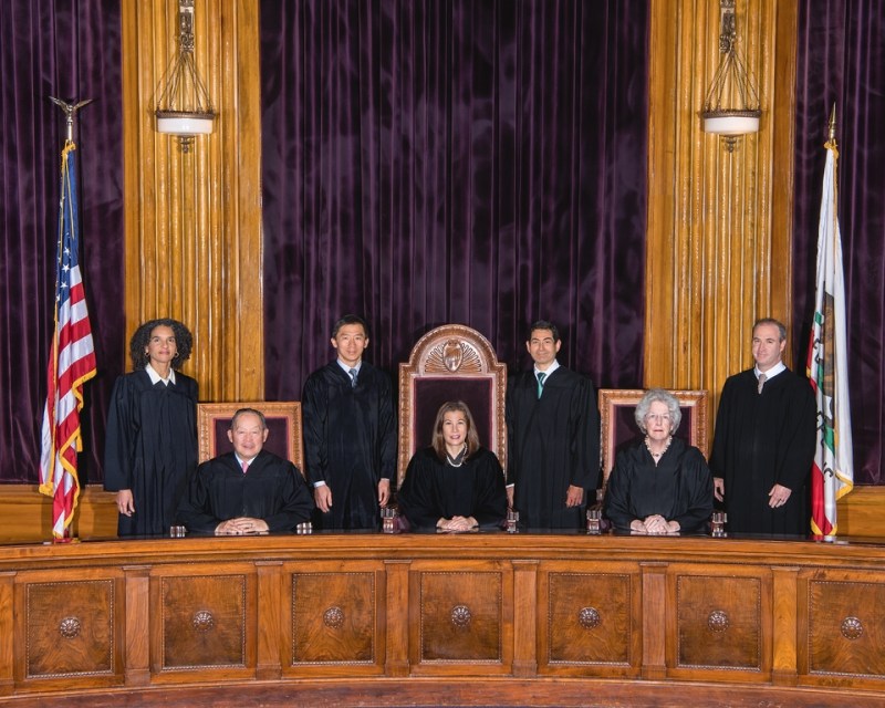 Seven judges in black robes pose behind a large wooden bench in a courtroom, with US and California flags visible on either side.