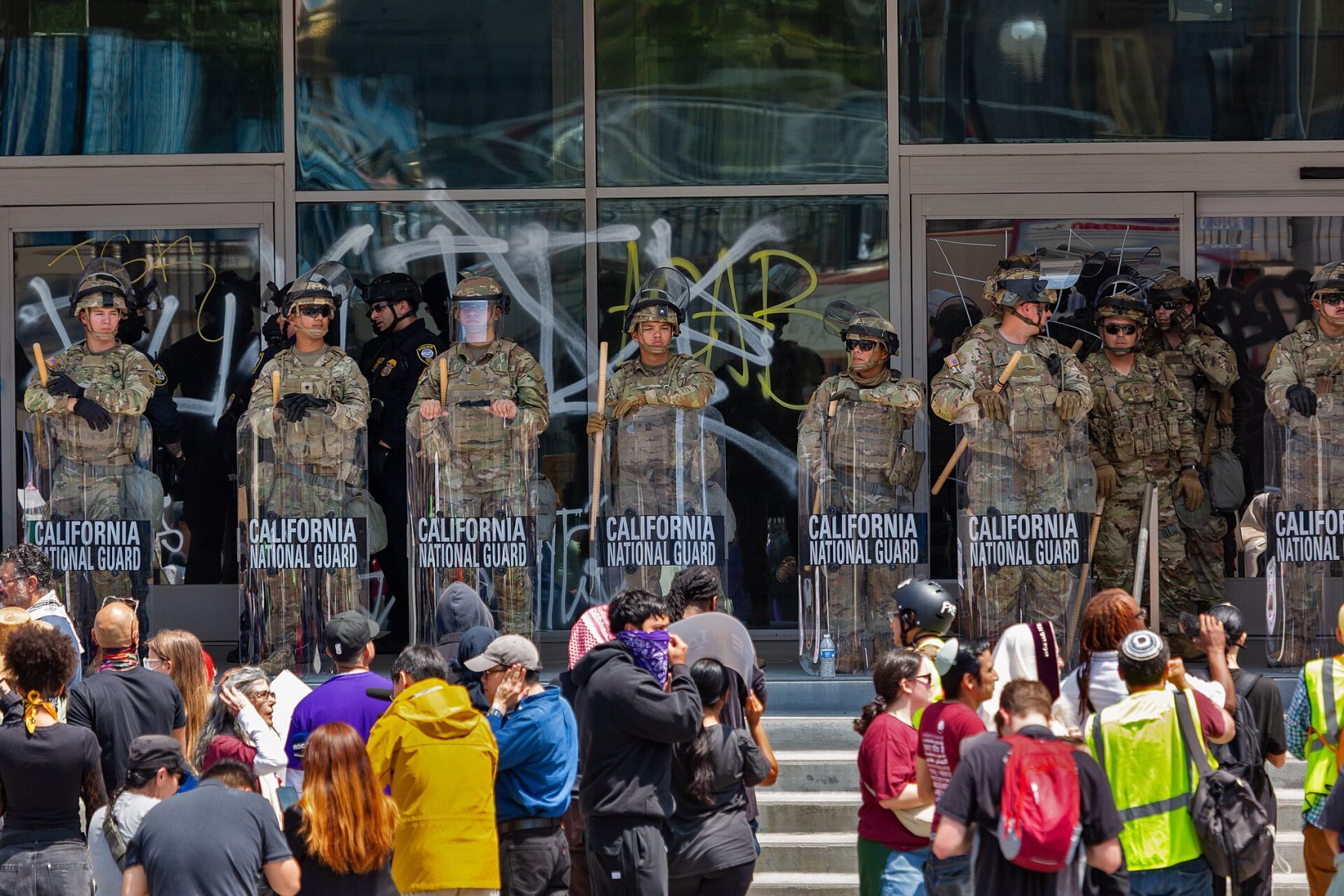 A line of National Guard troops stands in front of a building. They wear camo uniforms and armor, and carry batons and plexiglass shields that are marked "California National Guard." In front of them is a crowd of people, seen mostly from the back, wearing a variety of colors.