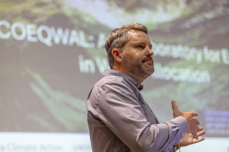 A man gesticulates with his hands while standing in front of a large screen.