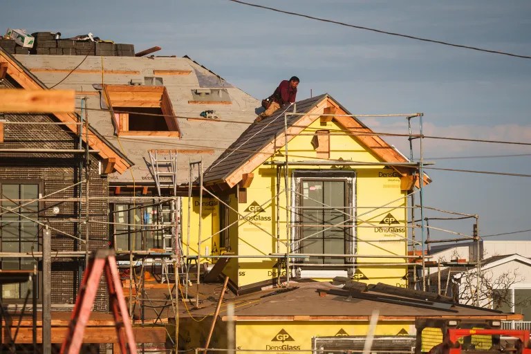 A worker stands on the roof of a bright yellow house under construction, surrounded by scaffolding and unfinished homes beneath a clear sky.