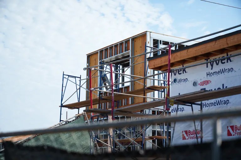 An unfinished multi-story building with exposed timber and steel framing rises against a pale blue sky; wrap-covered walls and scaffolding show active construction.