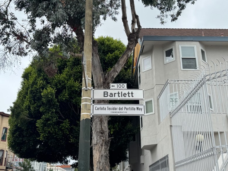 Street sign for Bartlett stands above a commemorative sign reading “Carlota Teixidor del Portillo Way.” A tree and a light gray building, located just steps away from Corey’s Pizza, are in the background.