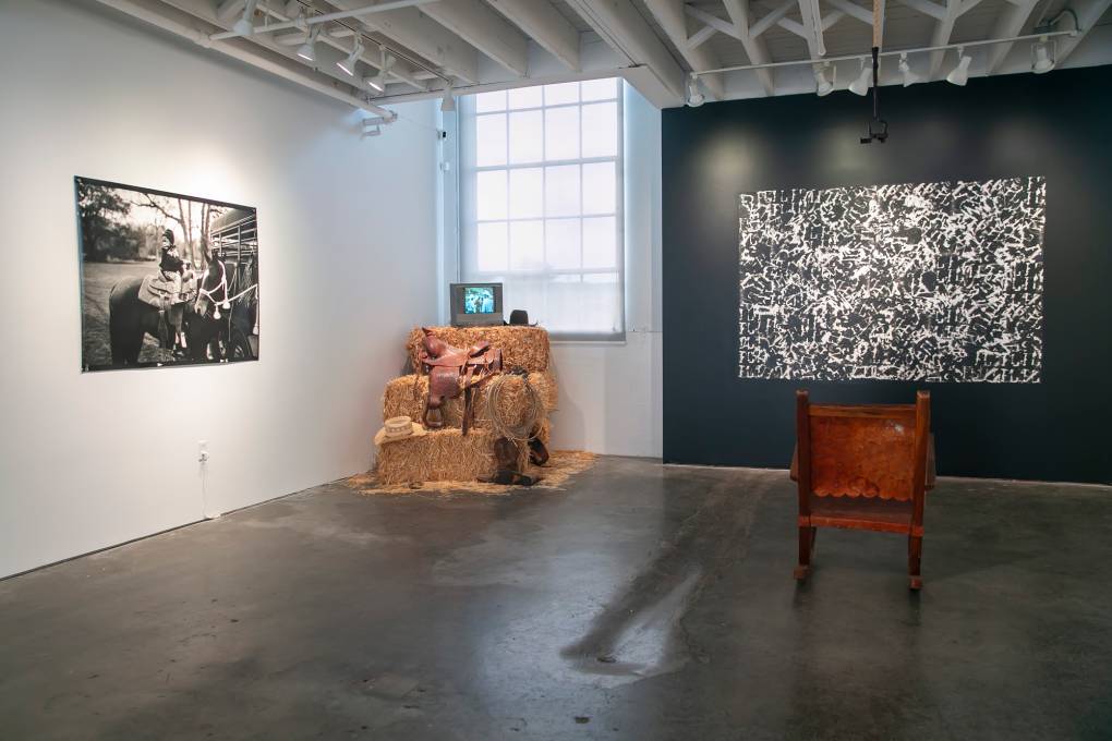Black and white photos on gallery walls, hay bales piled in corner with video monitor on top