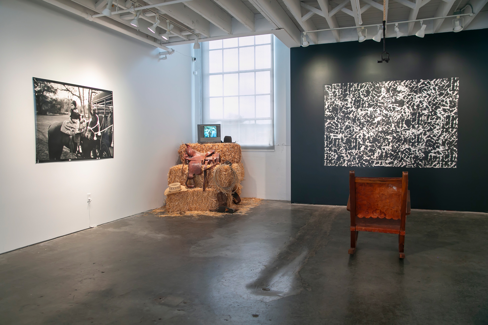 Black and white photos on gallery walls, hay bales piled in corner with video monitor on top
