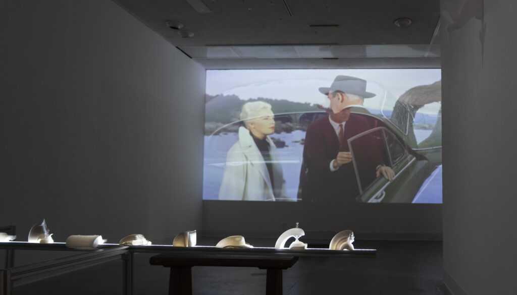 A dimly lit gallery room featuring a large video projection on the back wall showing a scene with two people near a classic car. In the foreground, a long, illuminated horizontal table displays a row of small, white 3D-printed sculptures.