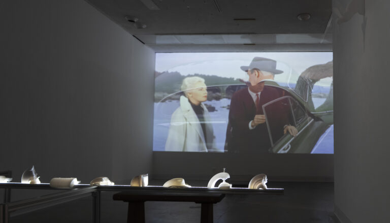A dimly lit gallery room featuring a large video projection on the back wall showing a scene with two people near a classic car. In the foreground, a long, illuminated horizontal table displays a row of small, white 3D-printed sculptures.