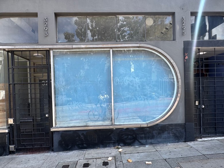 Gray building facade with two black doors, large rounded-corner window covered in blue film, and faint graffiti; debris is scattered on the sidewalk in front, hinting at the building’s past mission.