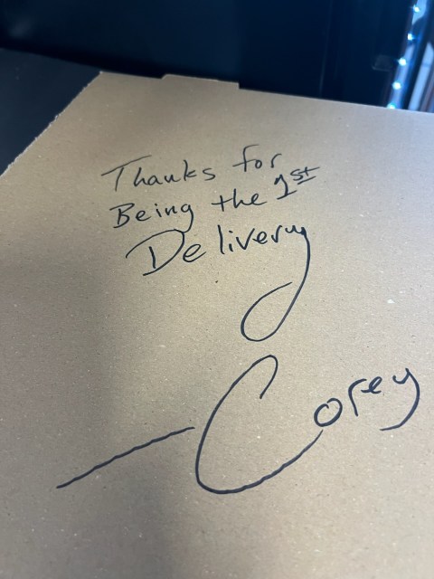 A handwritten note on cardboard reads, “Thanks for being the 1st Delivery. -Corey.” The background includes a black surface and some visible lights.