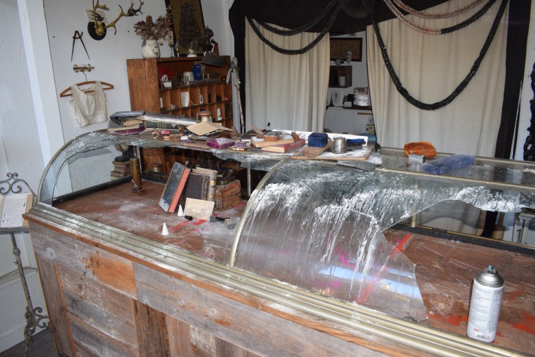 A damaged glass display counter in a cluttered shop, with scattered books, papers, and various objects. A spray paint can is on the counter. Shelves and curtains are visible in the background.