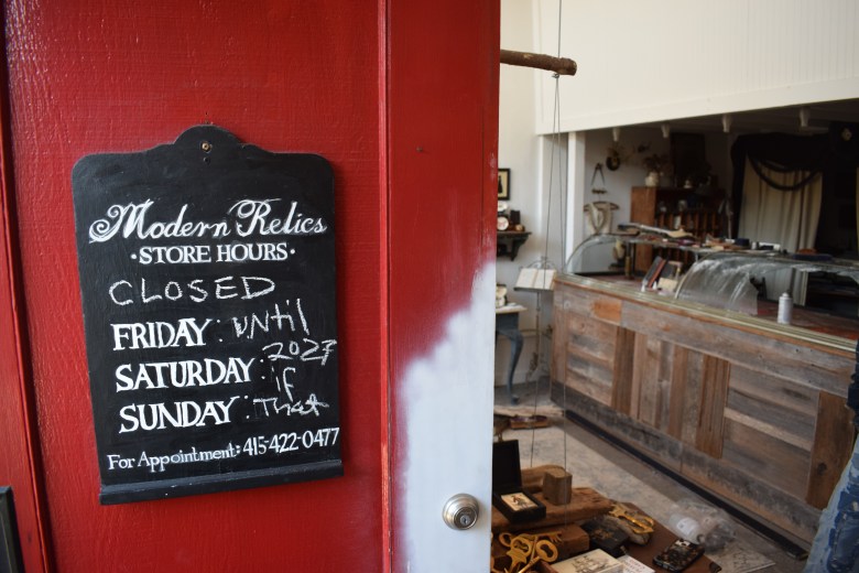 A red door with a sign reading “Modern Relics Store Hours: CLOSED” and unclear handwritten notes about opening times. The interior shows a cluttered counter and various vintage items.