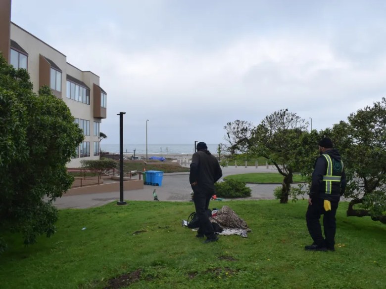 Two people in dark clothing stand on a grassy area near a building, facing toward a beach and the ocean under a cloudy sky.
