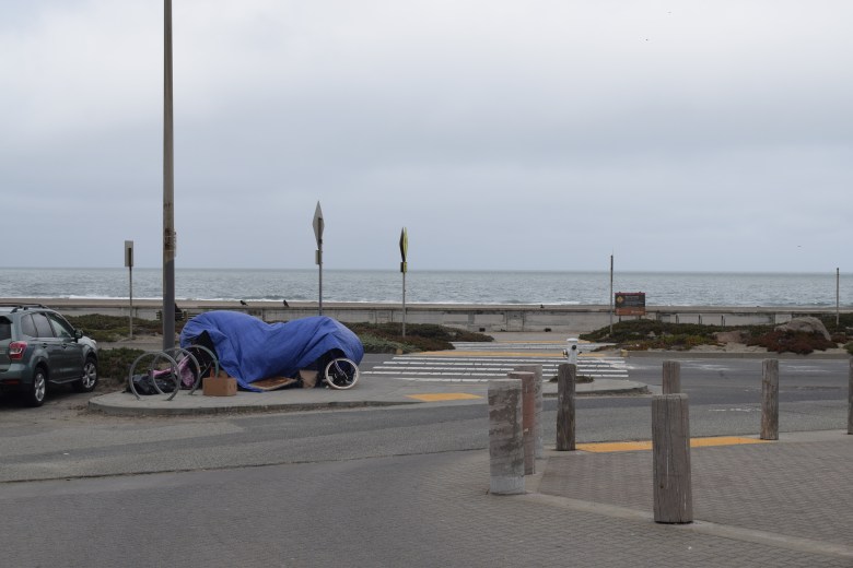 Bicycles and belongings covered by a blue tarp are parked by the street near the ocean, with cloudy skies and a few cars nearby.
