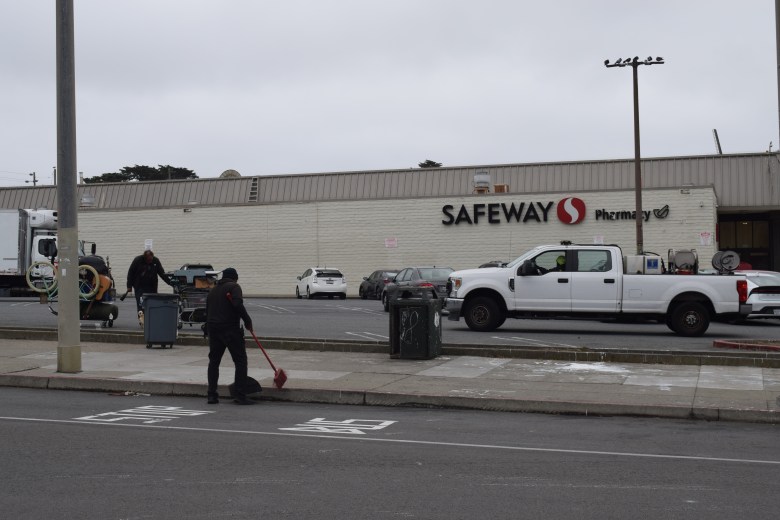A person sweeps the sidewalk in front of a Safeway store with a pharmacy; several vehicles are parked outside on a cloudy day.