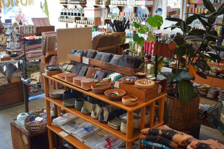 Display shelves in a store with ceramics, wallets, small accessories, potted plants, and various household goods arranged neatly in a well-lit space.