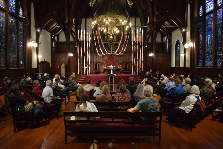 A group of people sit in pews facing a speaker at a podium in a large, ornate church with wooden beams and stained glass windows.