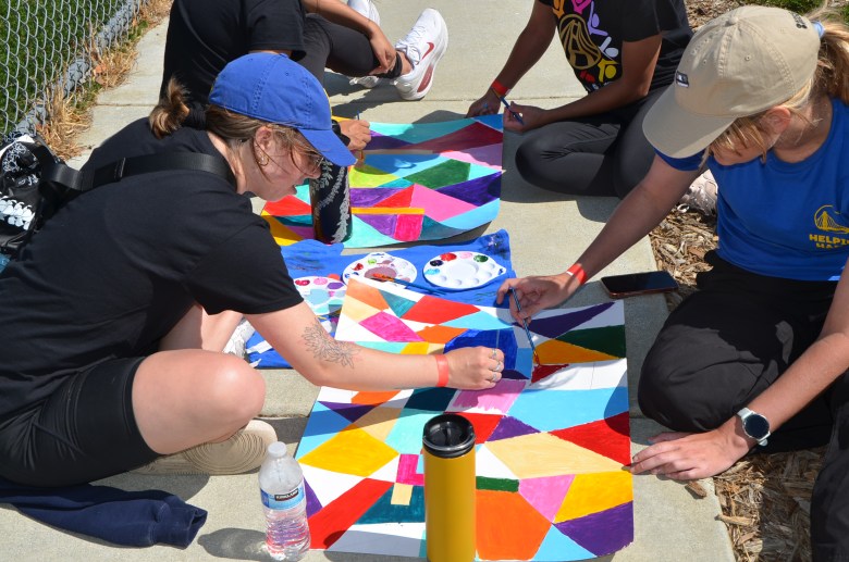 Four people sit on pavement outdoors like creative warriors, painting colorful geometric patterns on paper using paint palettes. Water bottles and cups are nearby.