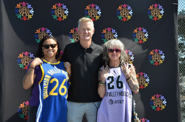 Three adults stand in front of a Warriors logo backdrop; the women on either side hold up basketball jerseys with the number 26, while the man in the middle smiles at the camera.