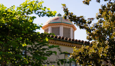 A decorative dome with floral patterns rises above a tiled roof, framed by lush green foliage against a clear blue sky.