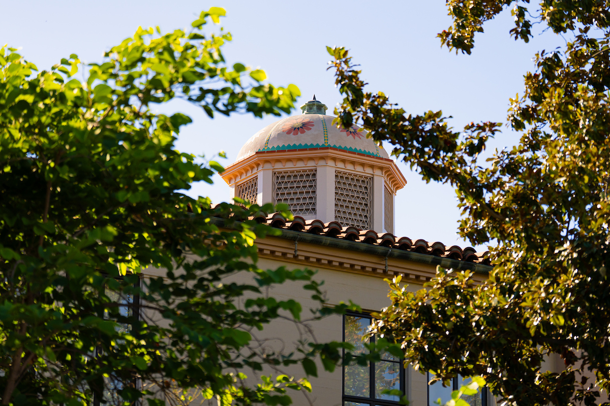 A decorative dome with floral patterns rises above a tiled roof, framed by lush green foliage against a clear blue sky.