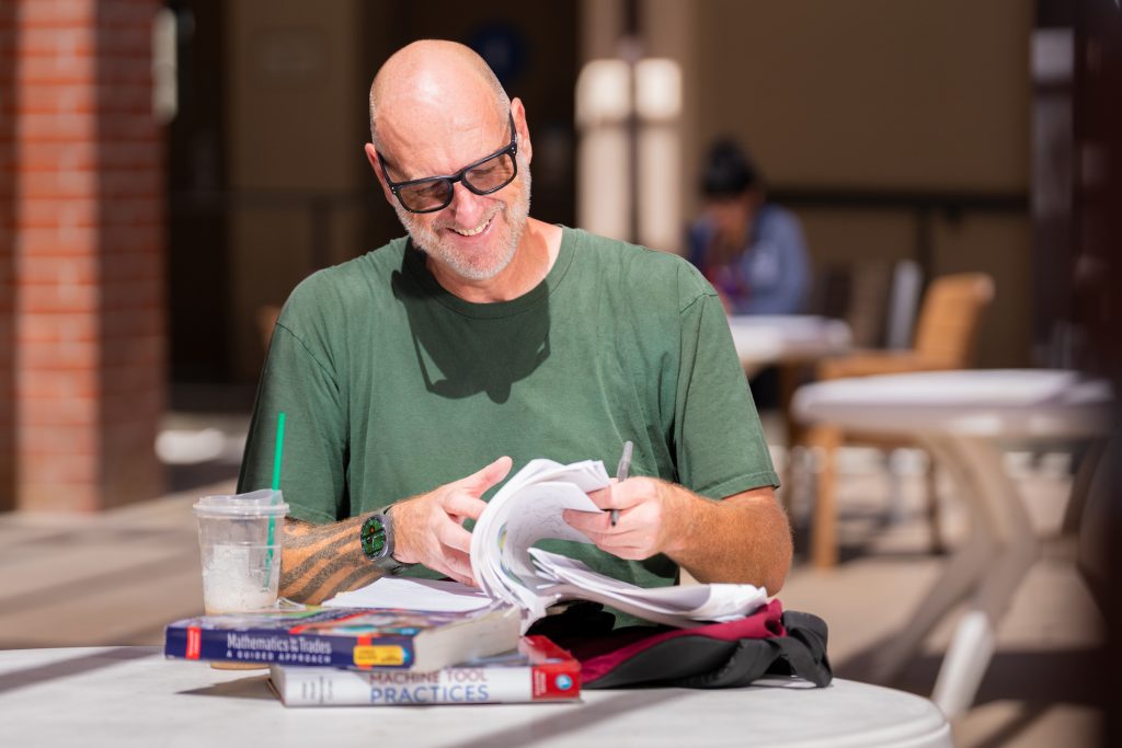 A person with glasses and a beard smiles while studying outdoors, surrounded by textbooks and a drink on a table.