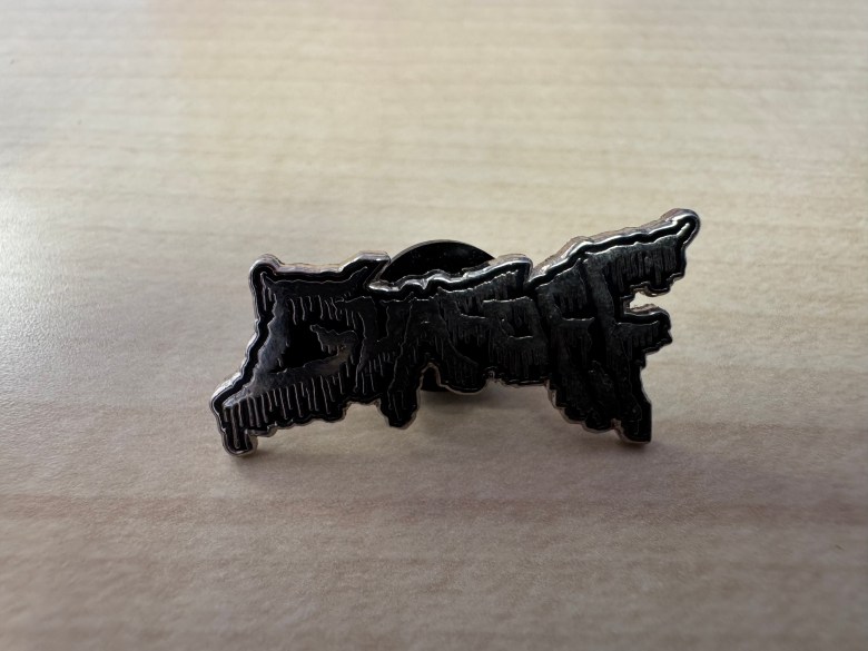 A black enamel pin with jagged edges and a dripping text design resting on a light wood surface.