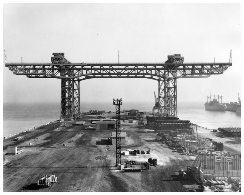 Large metal gantry structure under construction at a dockyard near the ocean, with equipment, supplies, and ships visible in the background.