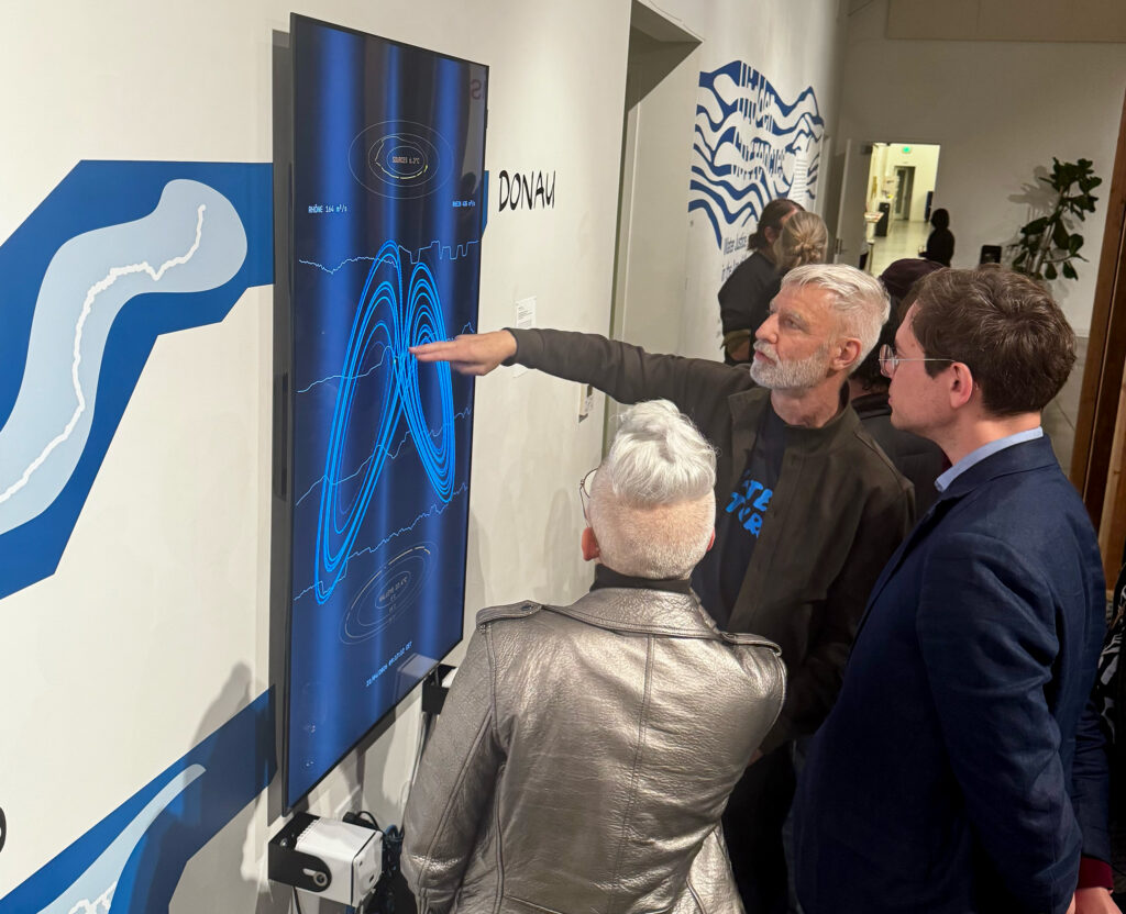 Greg Niemeyer pointing at a vertical digital display showing a blue mathematical graph on a gallery wall. Two visitors stand alongside him, watching the data visualization while blue river graphics surround the screen.