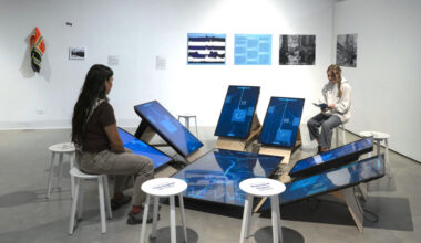 Two gallery visitors sit on white stools around a circular art installation by Greg Niemeyer, featuring multiple tilted digital screens displaying blue data visualizations on the floor of a white-walled gallery.