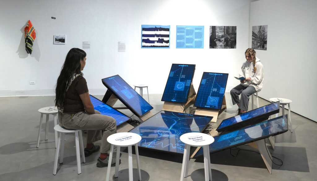 Two gallery visitors sit on white stools around a circular art installation by Greg Niemeyer, featuring multiple tilted digital screens displaying blue data visualizations on the floor of a white-walled gallery.