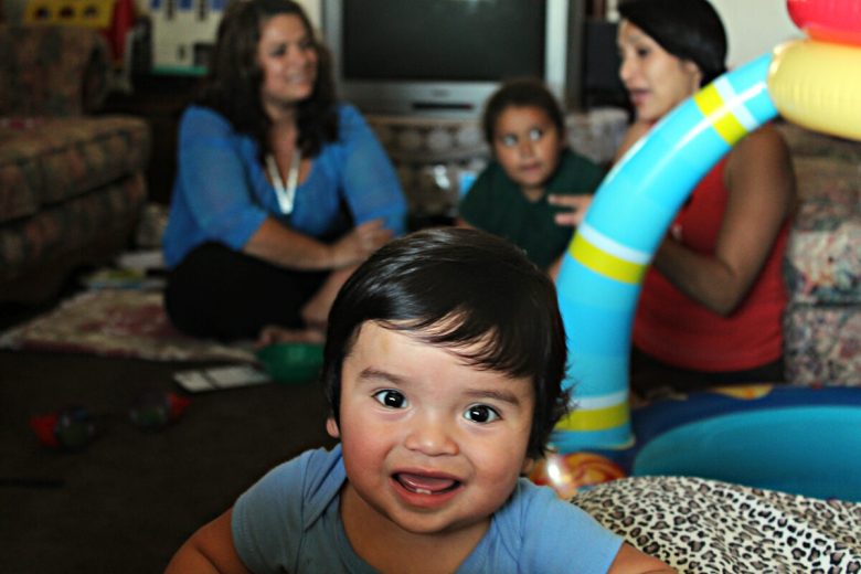 Smiling baby in a light blue shirt in front of family members and a large colorful inflatable pool toy.