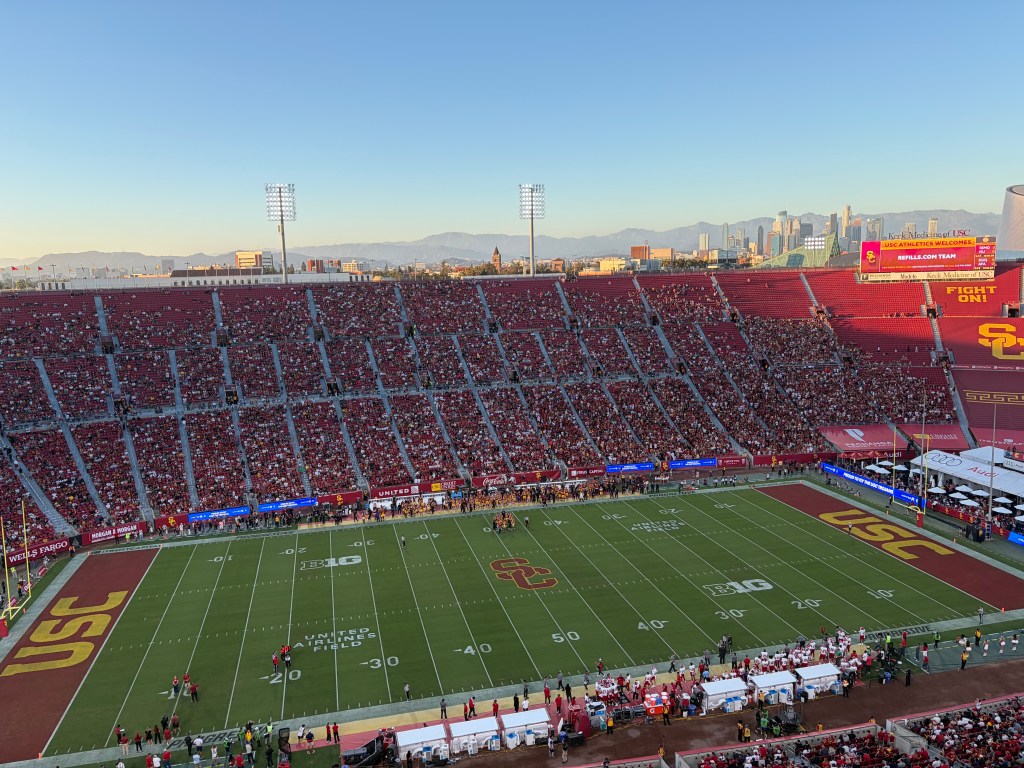 An aerial view of a football stadium with a game in progress between teams at United Airlines Field, with the downtown Los Angeles skyline in the background.