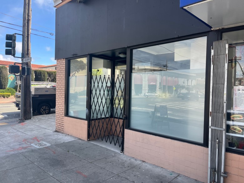 A storefront with large windows has black security gates partially closed across the entrance; the interior appears empty and a street intersection is visible outside.