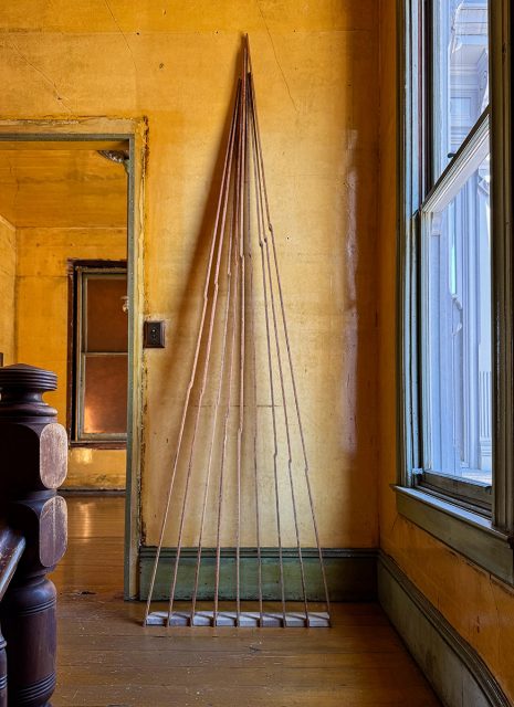 Six long wooden poles lean upright against a yellow wall in a sunlit, rustic room with wooden floors and large windows.