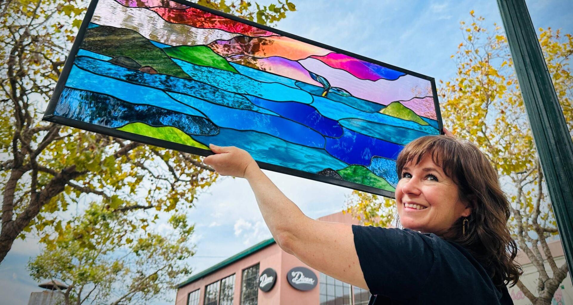 A woman in a black top turns and smiles at the camera while holding up a stained glass window showing the form of a sunset over the ocean