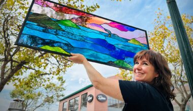 A woman in a black top turns and smiles at the camera while holding up a stained glass window showing the form of a sunset over the ocean