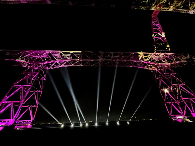 Steel bridge structure illuminated in purple at night, with bright white spotlights projecting upwards from the ground beneath its arch.
