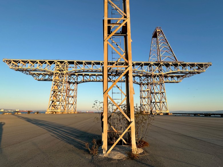 Large, rusted steel structure with a crossbeam stands on a wide, empty concrete lot under a clear blue sky, with some weeds growing at its base and long shadows cast by the setting sun.