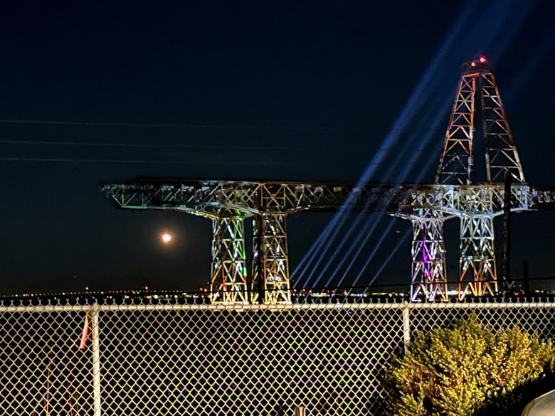 A large illuminated metal structure at night with beams of light shining upward, seen behind a chain-link fence and some plants.